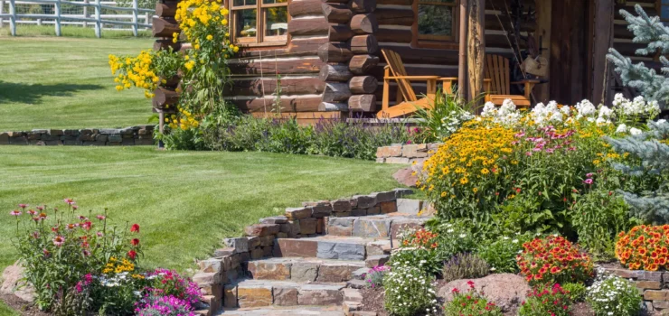 Stone steps lead to a log cabin with a porch, wooden chairs, and vibrant flower beds. The yard is lush and green, with a fence and hills in the background.